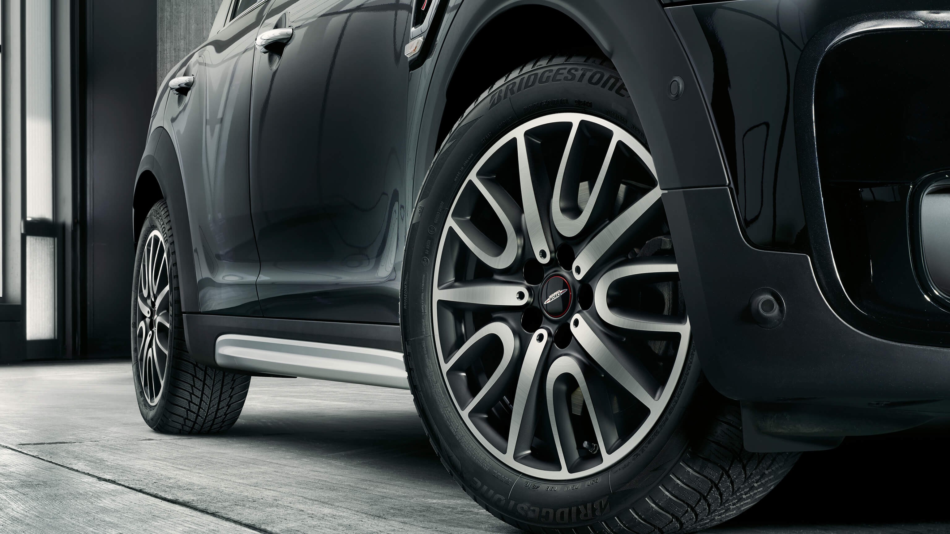 Close-up of a Mini Cooper Countryman wheel and tire, featuring stylish alloy rims and Bridgestone tires on a sleek black vehicle. The car is parked indoors on a concrete floor.
